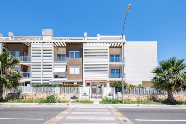 Un moderno edificio residencial de varios pisos con balcones, un acceso con portones y vista a la calle.