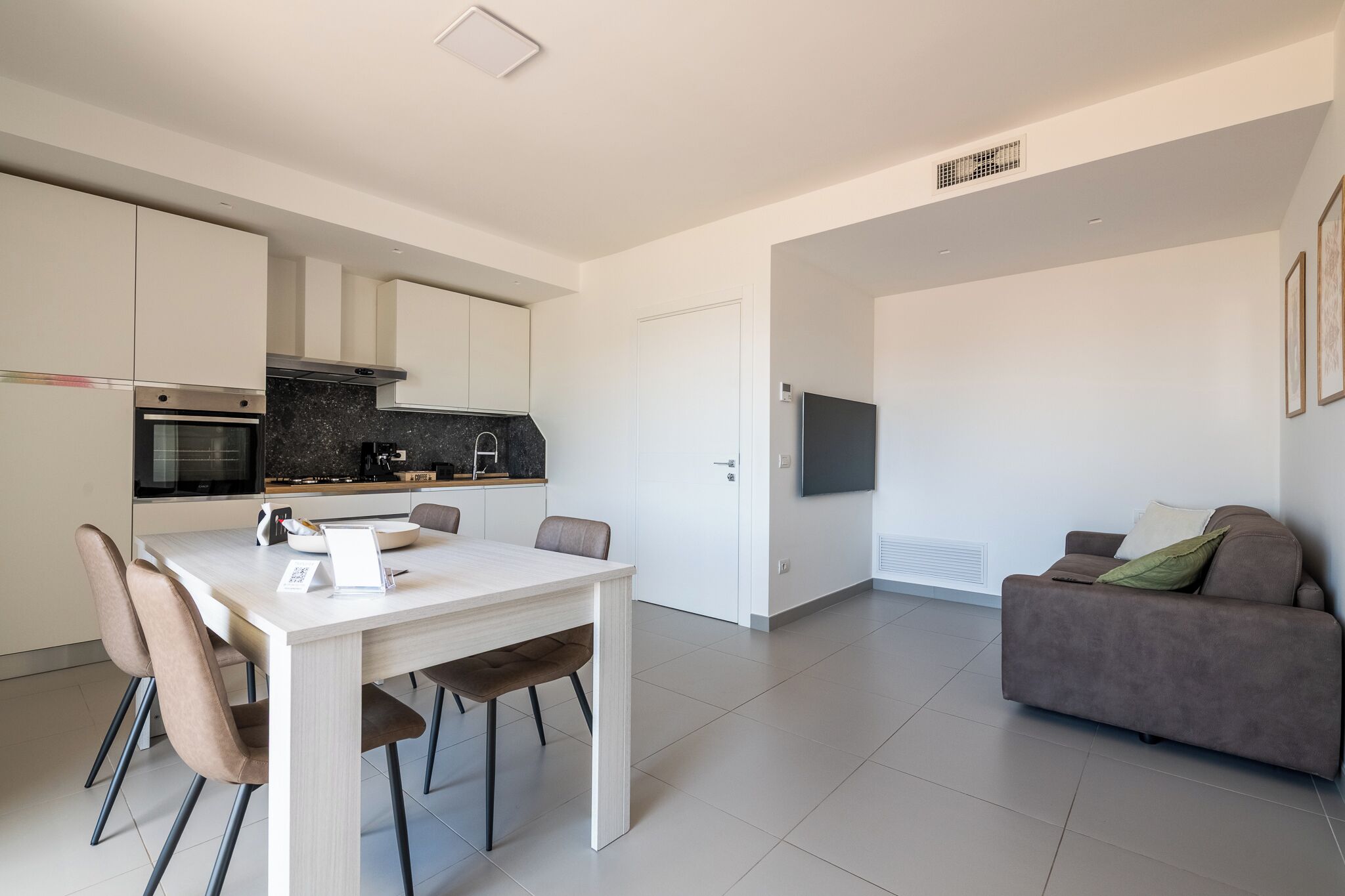 A modern kitchen with white cabinets, a dining table with chairs, and a grey sofa in a living area.