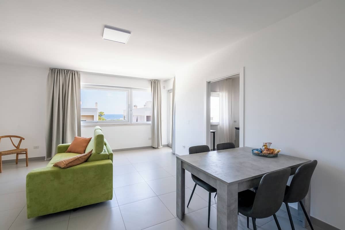 Un salón moderno con un sofá verde, una mesa de comedor con sillas y vistas al mar a través de amplias ventanas.
