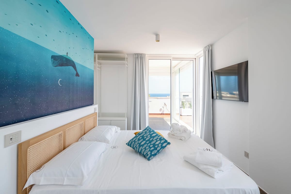 Habitación de hotel con una cama, un cuadro grande de una ballena, un televisor y un balcón con vistas.