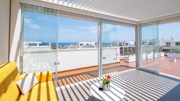 A modern balcony with a yellow sofa, a small table with a potted plant, and large glass doors offering a view of the city and the sea.