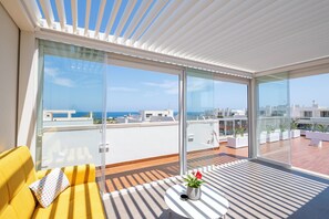 A modern balcony with a yellow sofa, a small table with a potted plant, and large glass doors offering a view of the city and the sea. - White Coast Relax & Stay (Polignano a Mare)