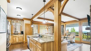 A spacious kitchen with wooden cabinets, a granite countertop, and a large island. There is a dining area with a table and chairs, and a view of the outdoors through large windows. - Chalakee Lodge CJ-3- Mountain View, Gated Resort, Indoor/Outdoor Pools & Hot Tub (Boone)