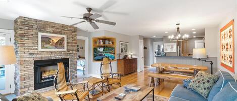 A living room with a stone fireplace, wooden furniture, and a view into the kitchen.