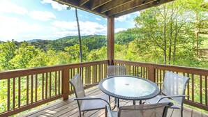 A wooden deck with a glass table and chairs, overlooking a forested landscape.