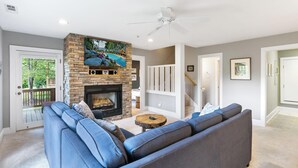 A living room with a stone fireplace, a ceiling fan, and a large window overlooking a deck.