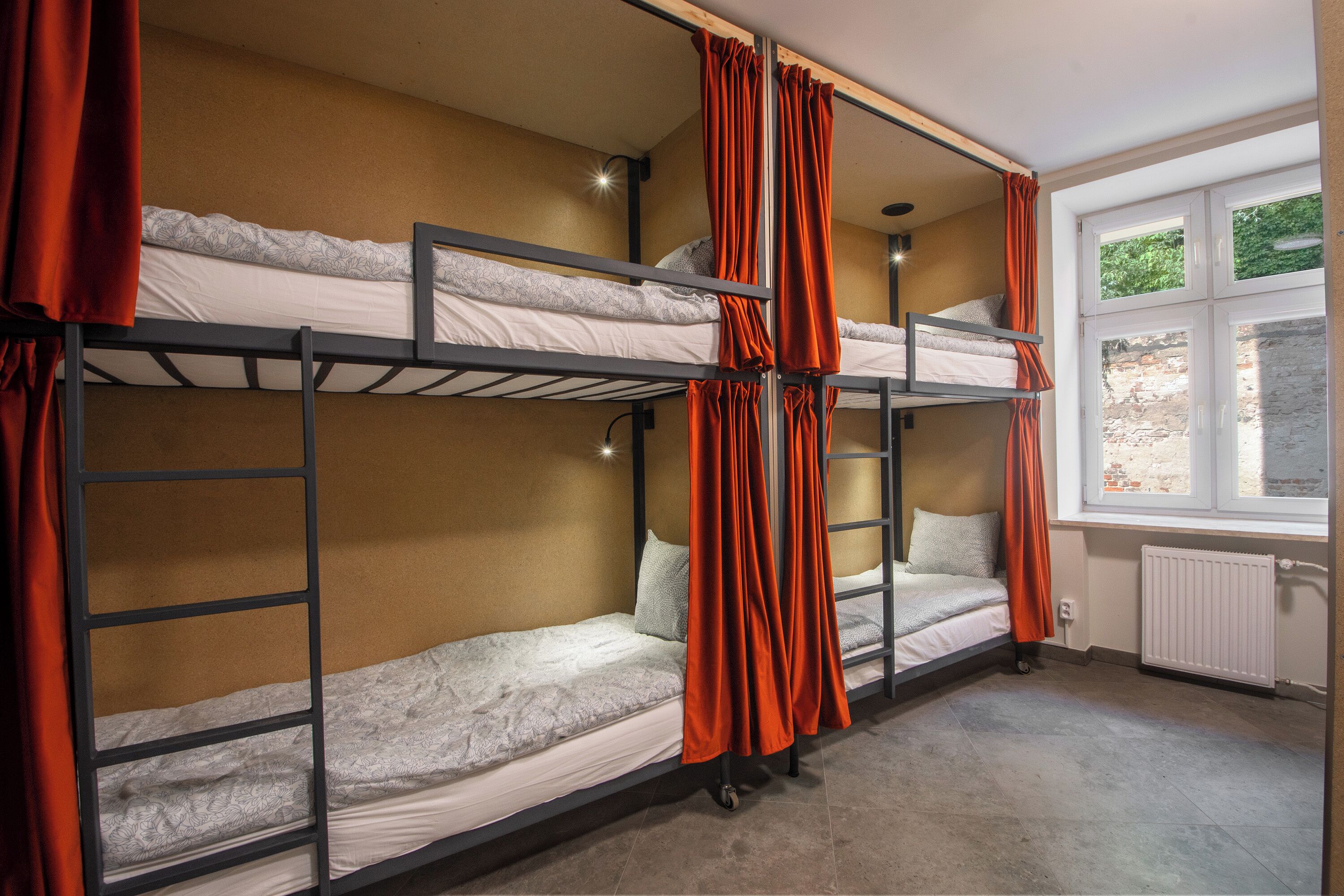 A dormitory room with bunk beds, a window with a view of greenery, and a radiator.