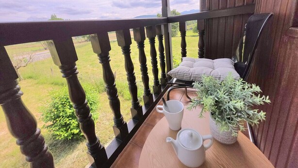 Un balcon en bois offrant une vue sur un champ verdoyant et des montagnes. On y trouve une table avec une tasse blanche, une théière blanche et une plante en pot.