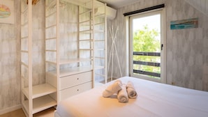 A bedroom with a large bed, a wardrobe with multiple shelves, a window with a view of greenery, and a painting on the wall.