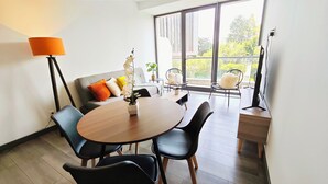 A modern living room with a round wooden table, black chairs, a grey sofa with colorful cushions, a floor lamp, and a large window with a view of greenery.