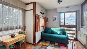 Una sala de estar acogedora con un sofá color turquesa, una mesa de comedor de madera con sillas, un tapete colorido y ventanas amplias que ofrecen vistas al exterior.