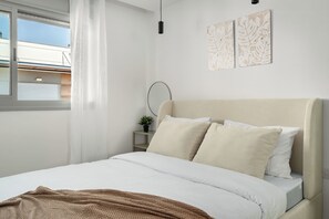 A bedroom with a bed, a window with curtains, a bedside table with a plant, and wall art.