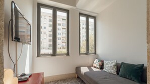 Quarto com sofá, televisão na parede e duas janelas com vista para um edifício.