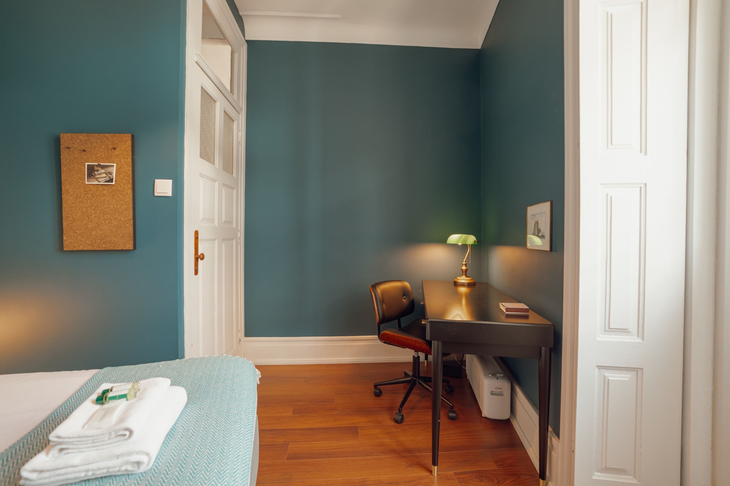 A room with teal walls, a bed with a blue and white bedspread, a desk with a lamp, and a chair.