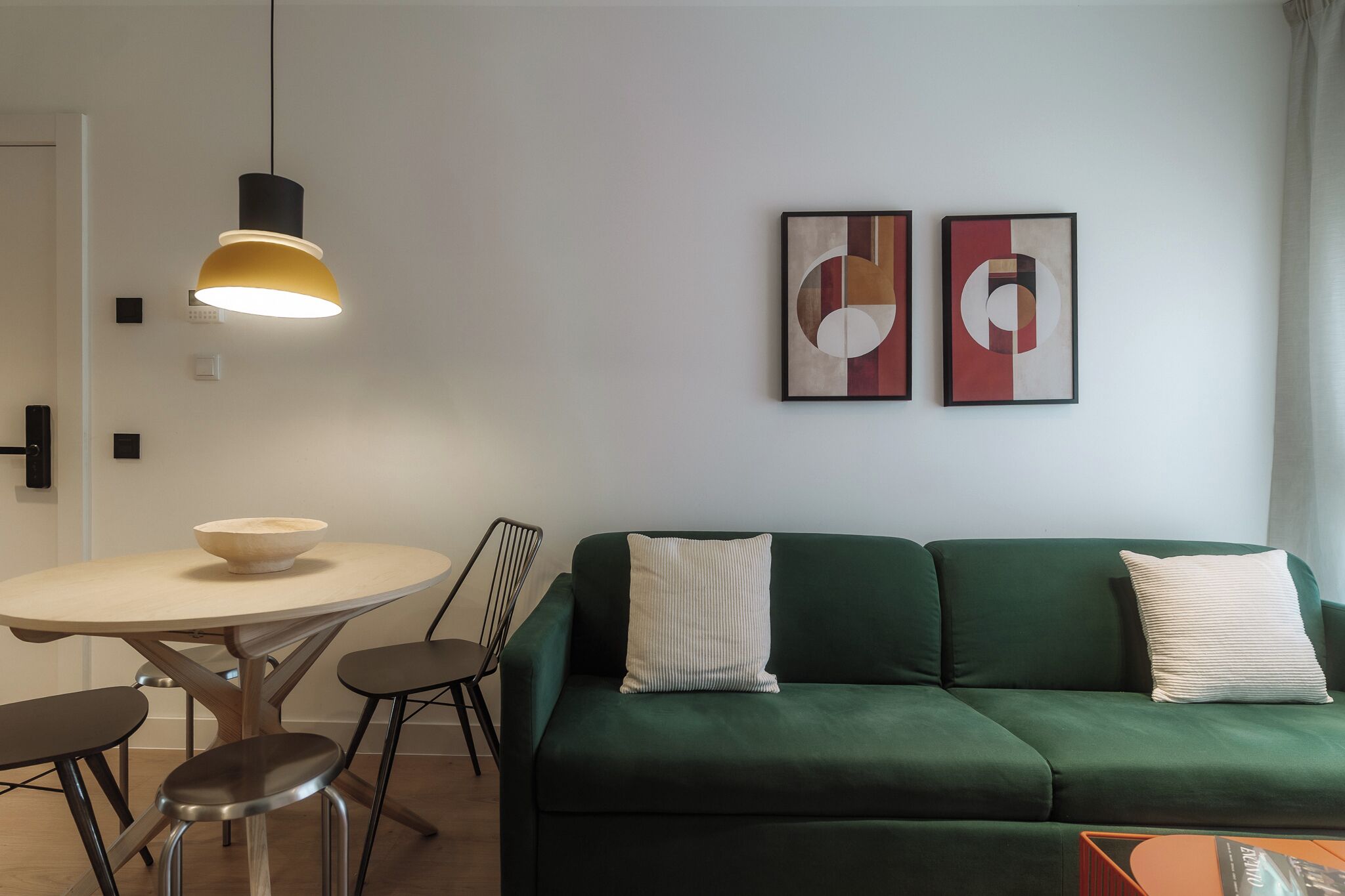 A modern living room with a green sofa, a round dining table with stools, a hanging pendant light, and two abstract paintings on the wall.