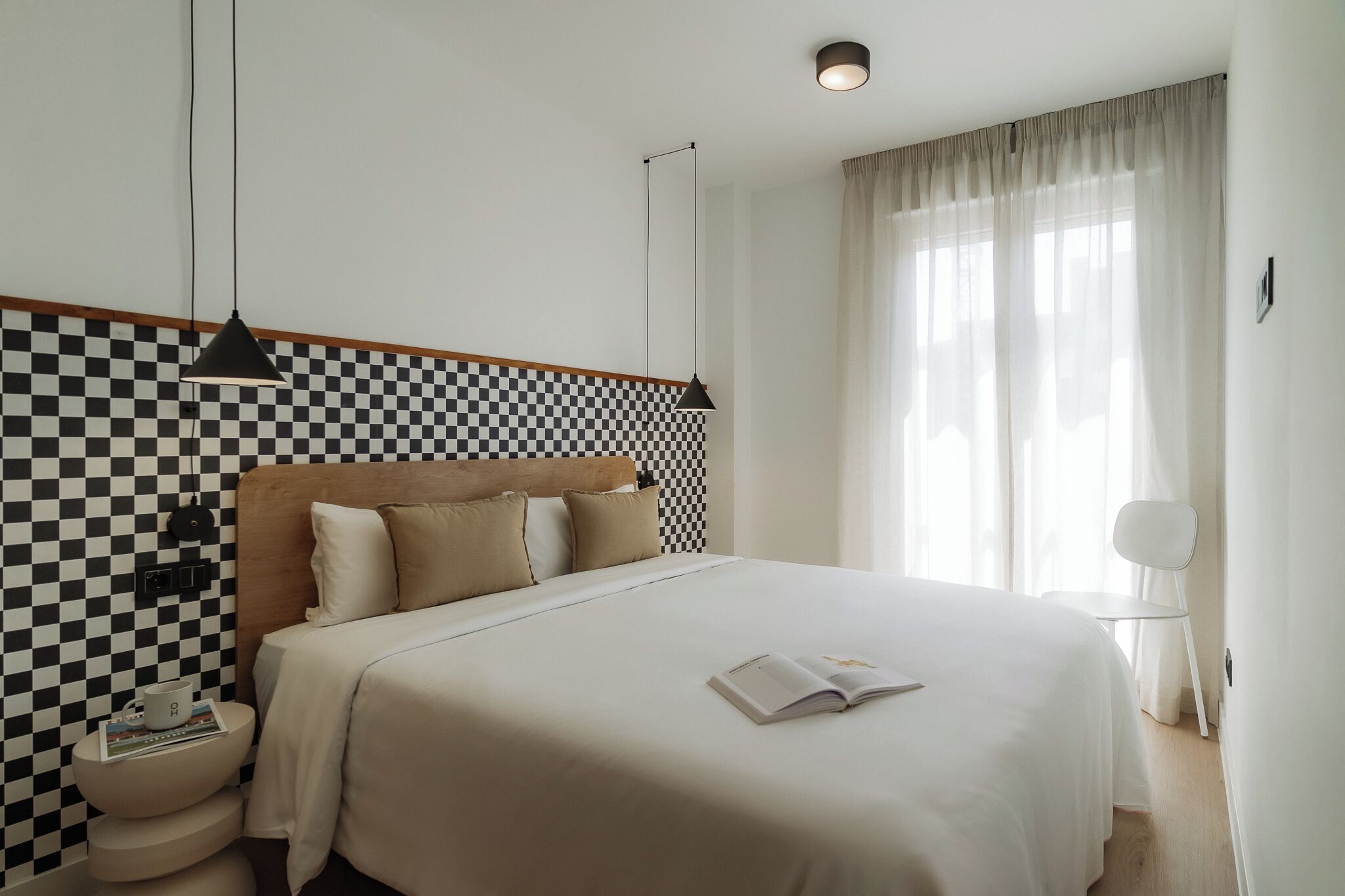 Un dormitorio ordenado con pared a cuadros, una cama con cubrecama blanco, una mesita de noche con un libro y una taza, y una lámpara colgante.