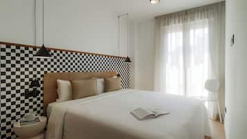 Un dormitorio ordenado con pared a cuadros, una cama con cubrecama blanco, una mesita de noche con un libro y una taza, y una lámpara colgante.