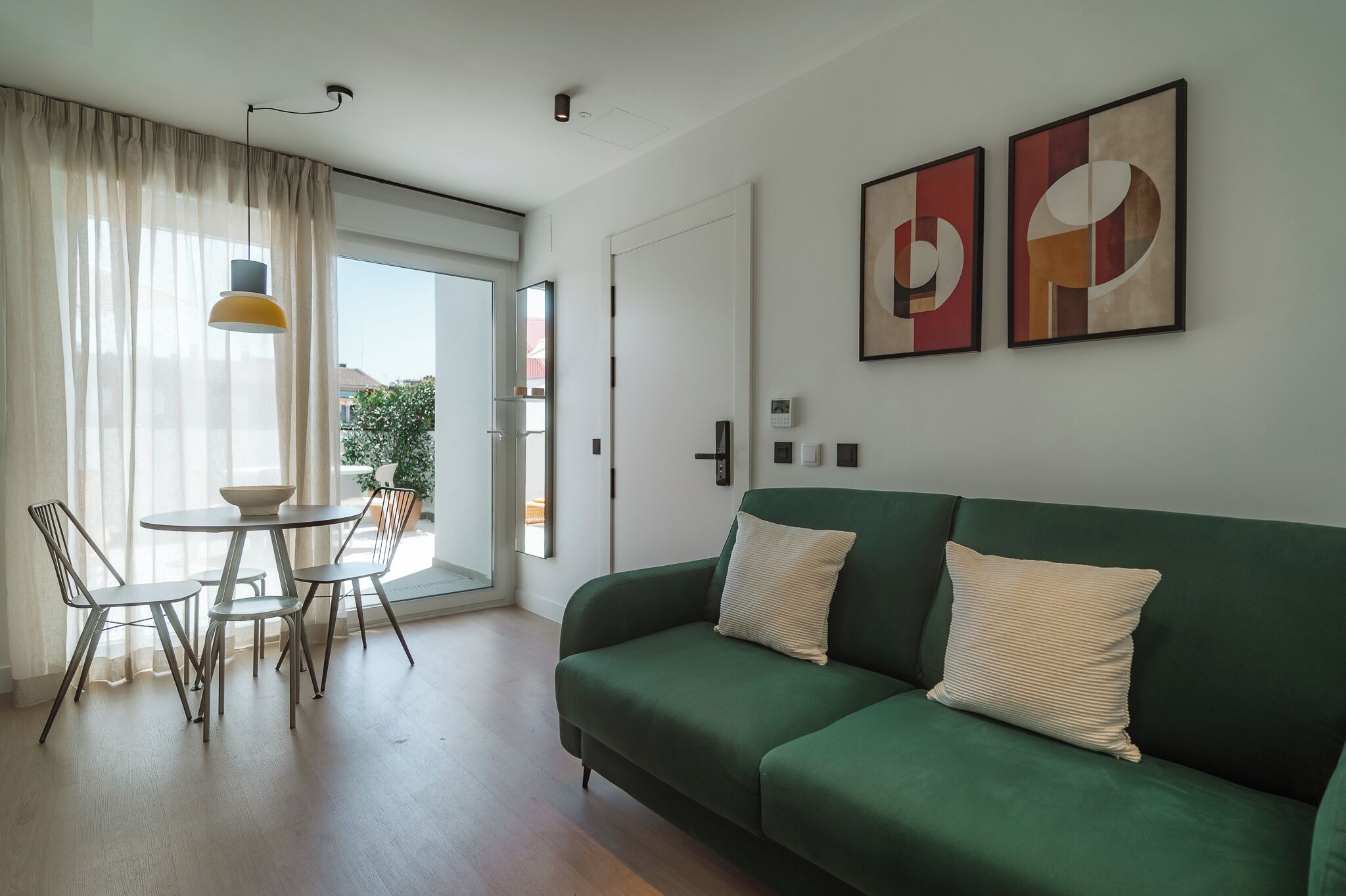 A modern living room with a green sofa, a round dining table with chairs, and a large window with sheer curtains.