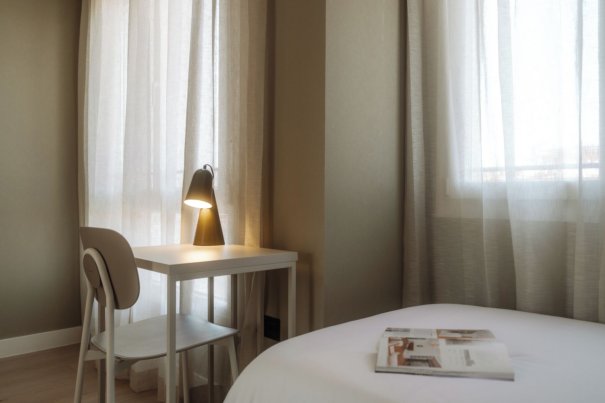 A hotel room with a bed, a desk with a lamp, a chair, and sheer curtains.