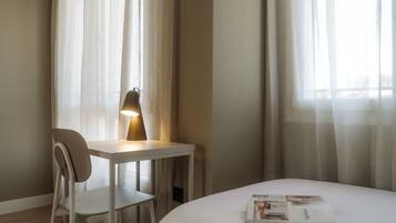 A hotel room with a bed, a desk with a lamp, a chair, and sheer curtains.