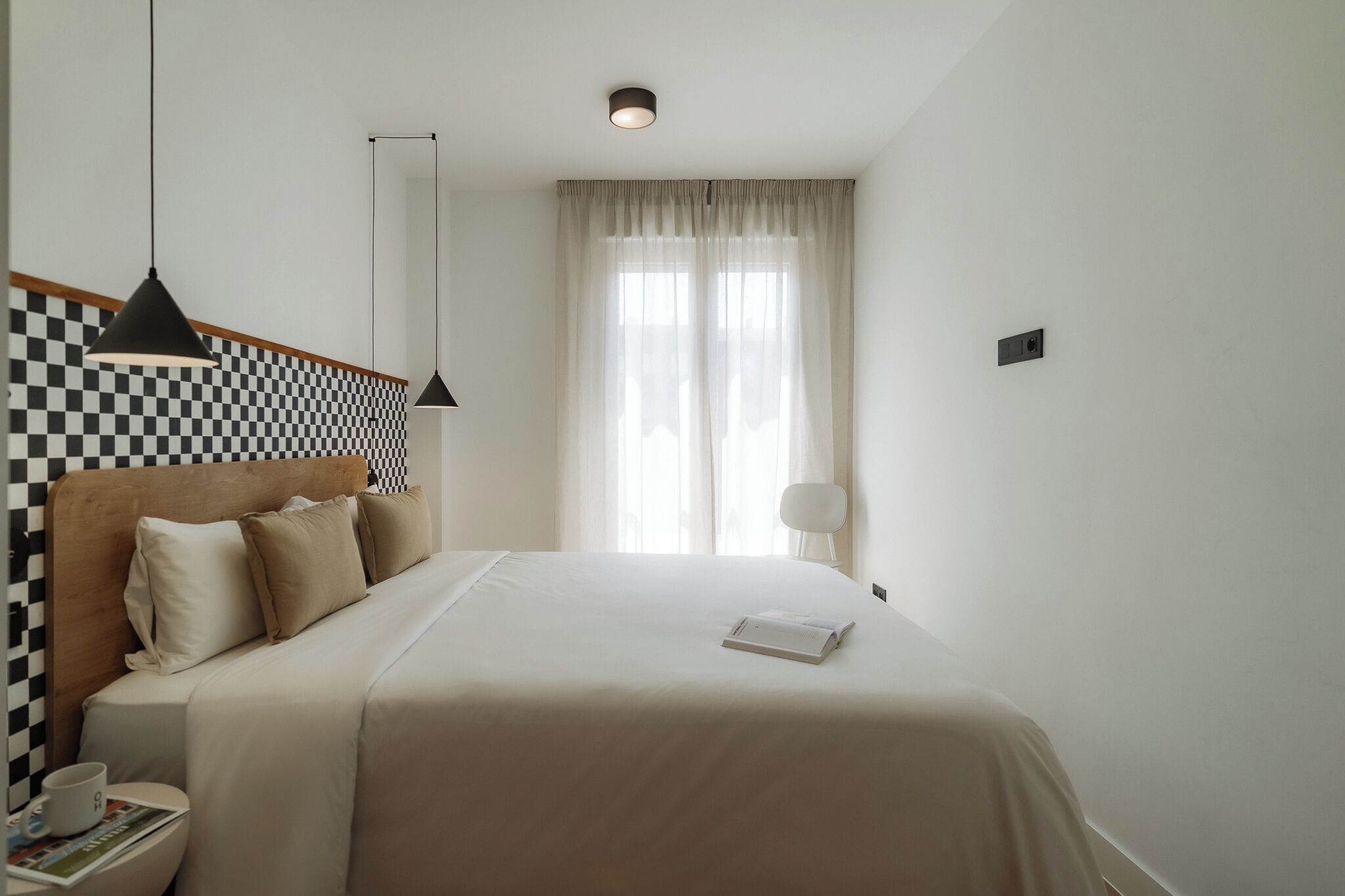 A neatly arranged hotel room with a checkered headboard, a large bed, a bedside table with a cup and magazine, and a window with sheer curtains.