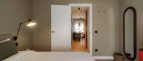 A hotel room with a bed, bedside lamp, and a mirror. There is a door leading to a dining area with a table and chairs.