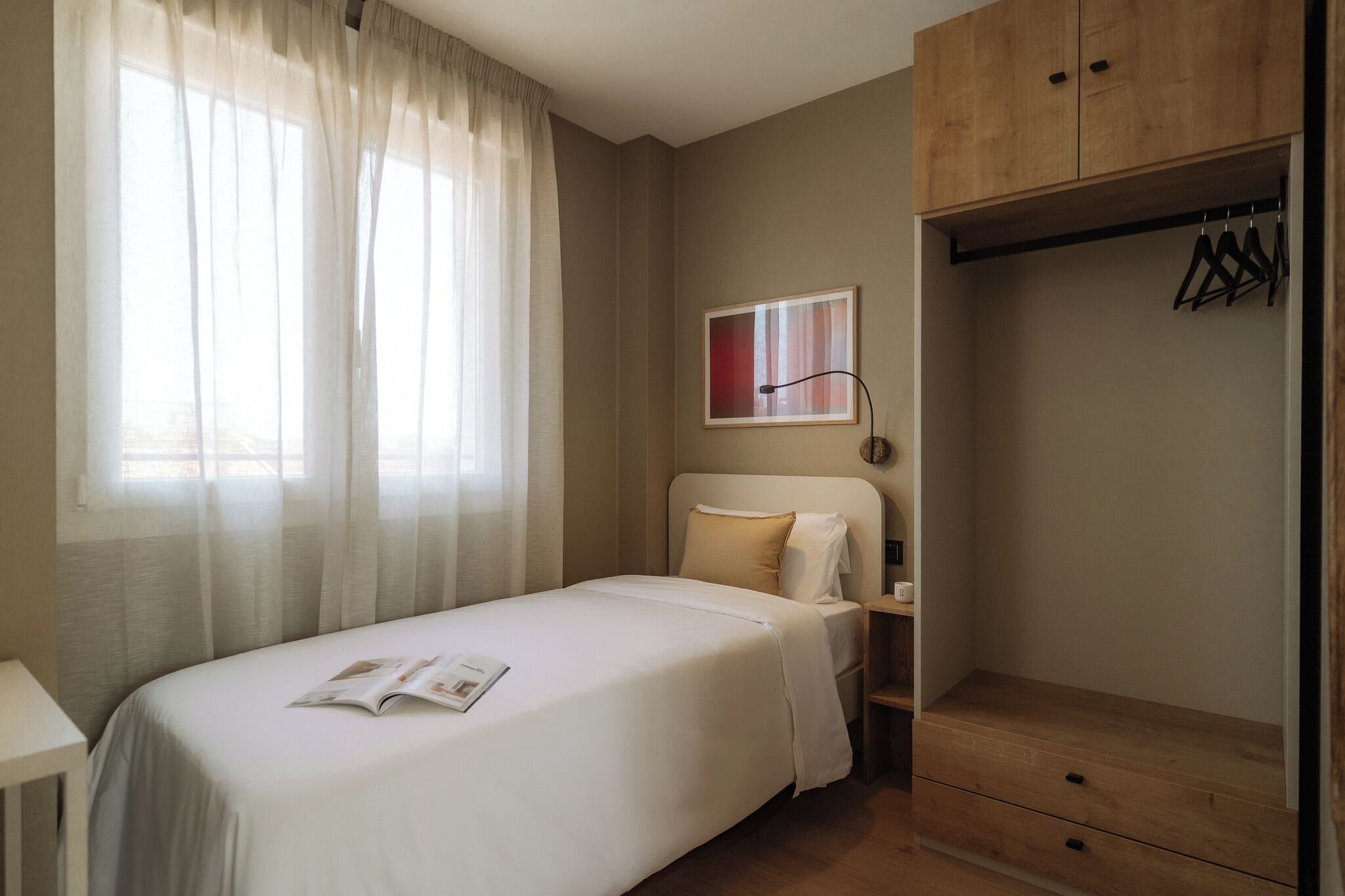 Un dormitorio ordenado con cama, mesita de noche, armario y ventana con cortinas translúcidas.