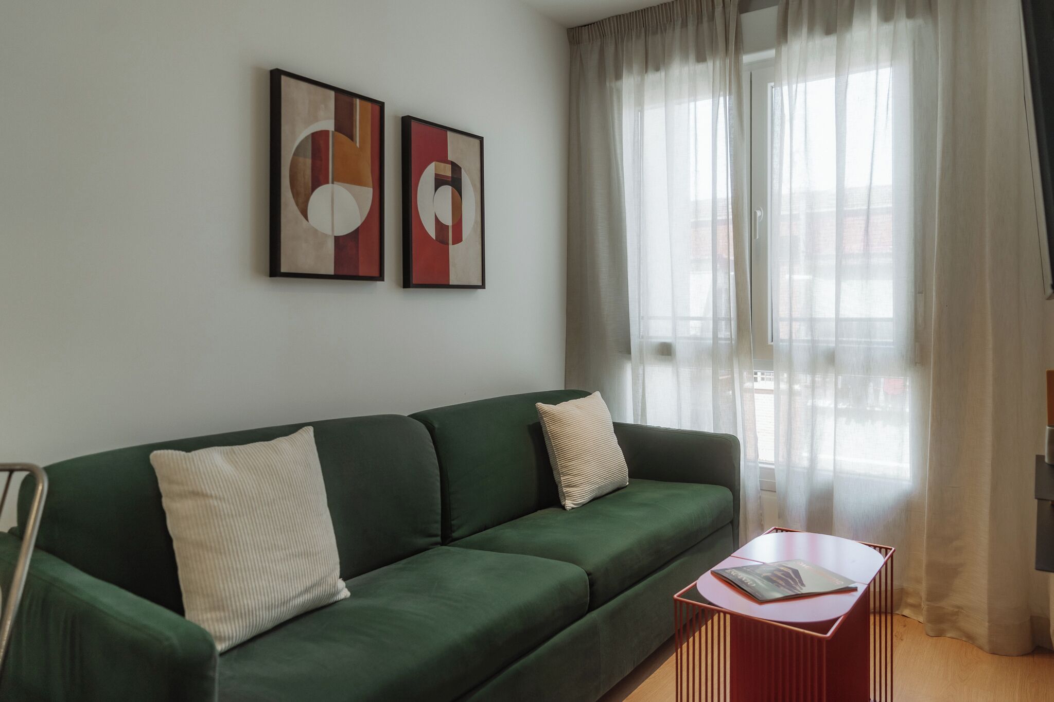Una sala de estar moderna con un sofá verde, dos cojines, una mesita de noche roja con una revista y arte abstracto en la pared.