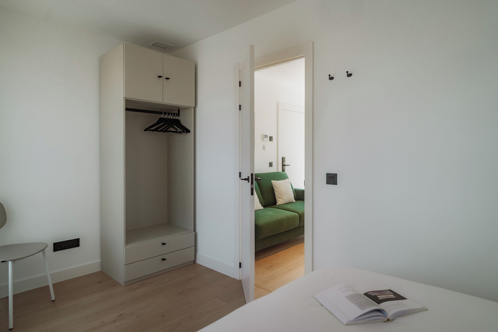 Un dormitorio minimalista con vestidor, un sofá verde en la habitación contigua y una mesita de noche con un libro.