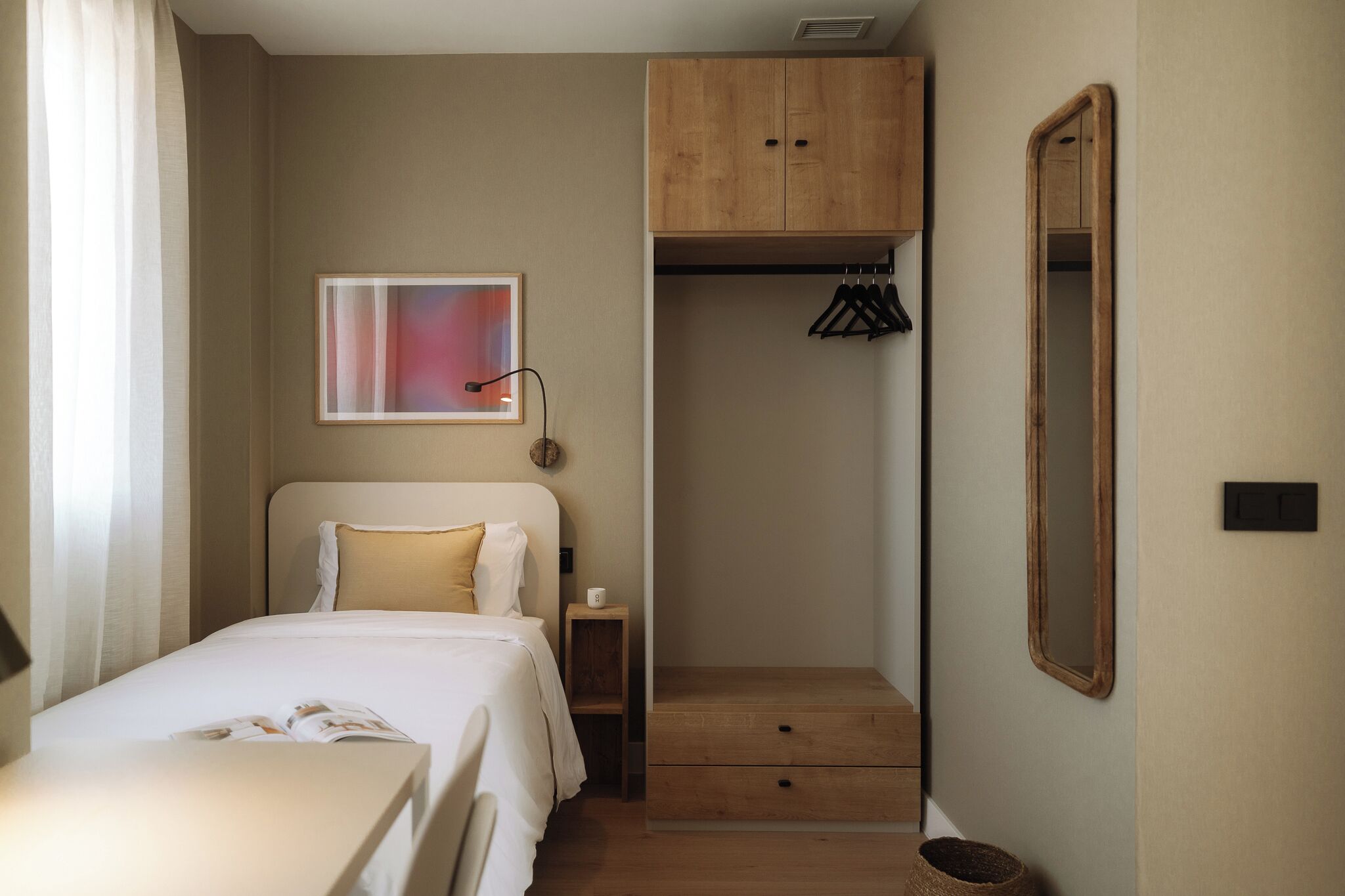 Hotellihuoneessa on sänky, yöpöytä, pieni kaappi, seinälle ripustettu naulakko ja peili.