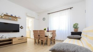 Un soggiorno moderno e luminoso con una TV a schermo piatto, mobili in legno, un'area pranzo con tavolo e sedie, un letto con lenzuola a righe e una pianta in vaso.