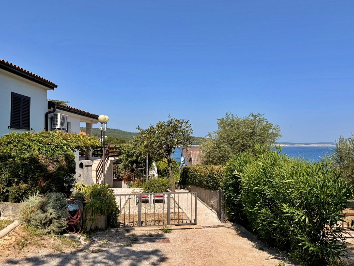 Una propiedad costera con un camino de entrada pavimentado que conduce a un acceso con puerta, rodeada de vegetación exuberante y vistas al mar.