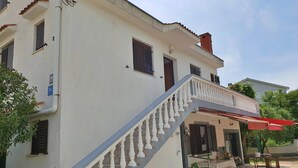 Un edificio de dos pisos con fachada blanca, una escalera con barandilla y un área de estar al aire libre cubierta.