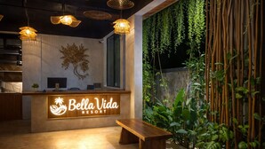 Un espace d’accueil avec un bureau en bois, un banc, des lampes suspendues en osier et un mur recouvert de plantes suspendues.