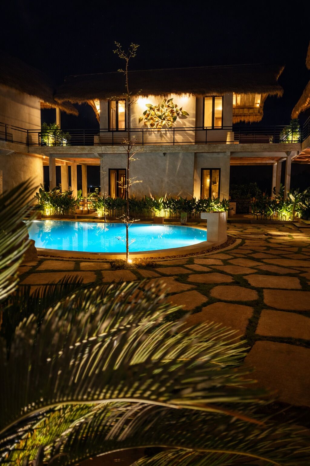 Un área de piscina con una construcción de techo de paja, suelo de piedra y plantas bien iluminadas.