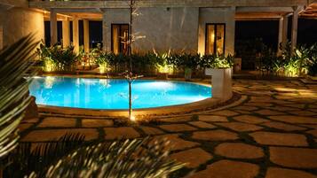 Un área de piscina con una construcción de techo de paja, suelo de piedra y plantas bien iluminadas.