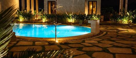 A pool area with a thatched-roof building, stone flooring, and well-lit plants.