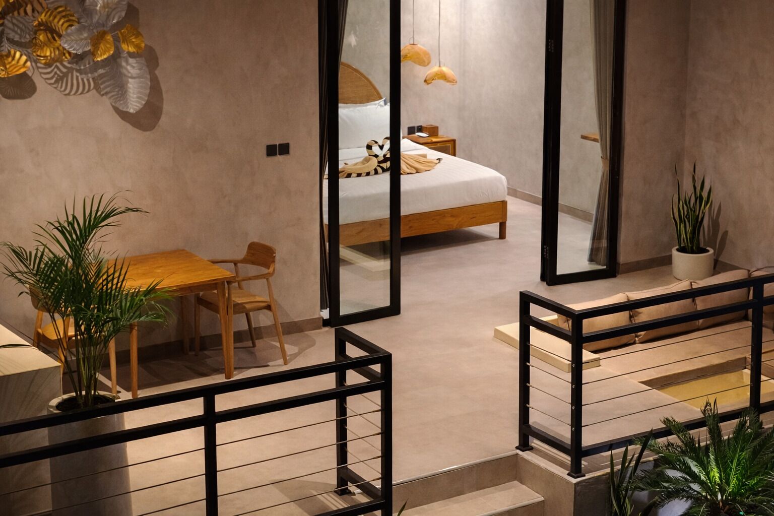 A modern bedroom with a bed, bedside table, and a small table with chairs. There are potted plants and a large mirror reflecting the room.