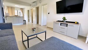 Een moderne woonkamer met een flatscreen-tv, grijze banken, een salontafel en een minimalistische kitchenette.