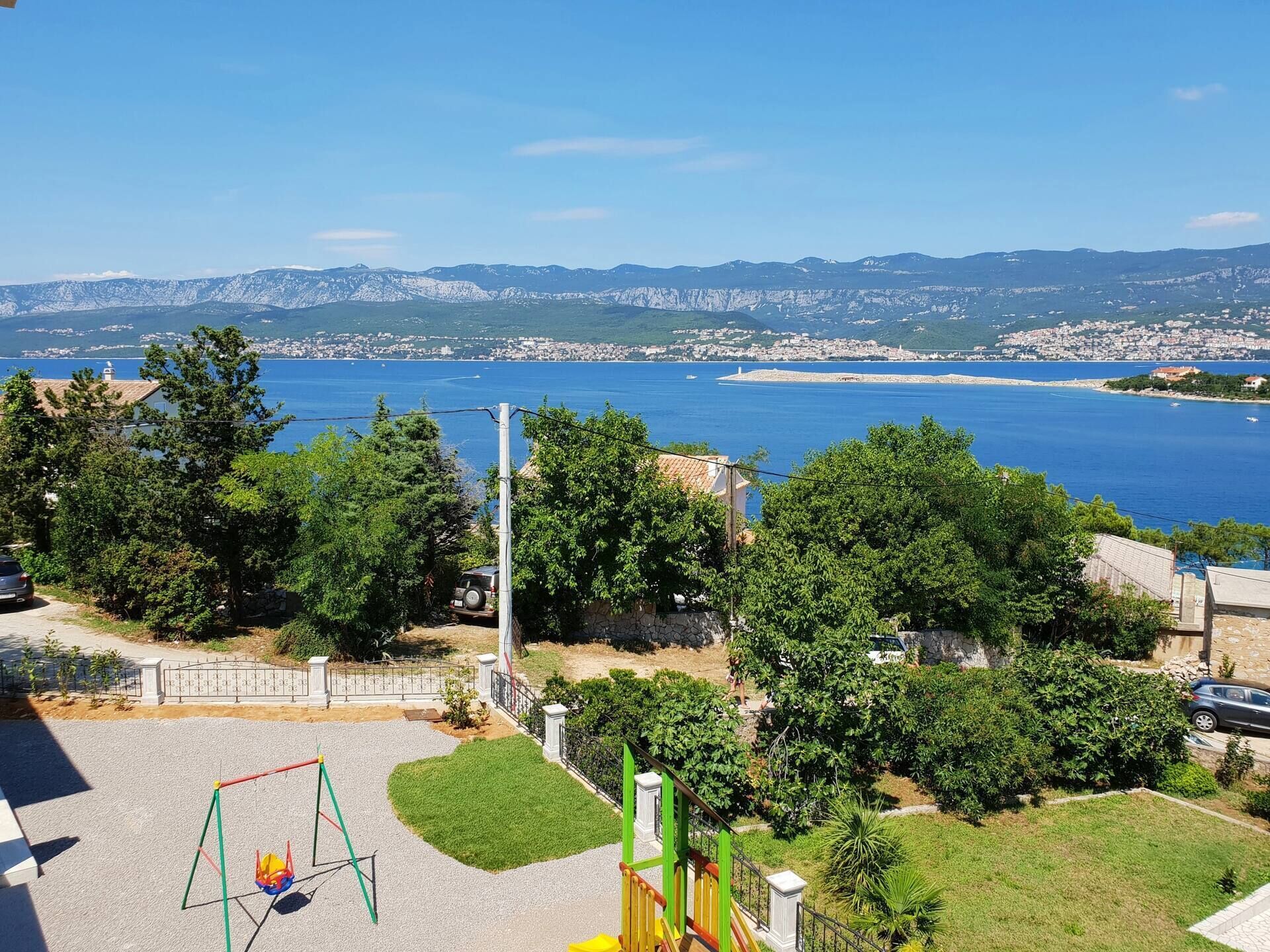 Ein Spielplatz mit Schaukeln und Rutsche, umgeben von viel Grün und Blick auf eine Küstenstadt.