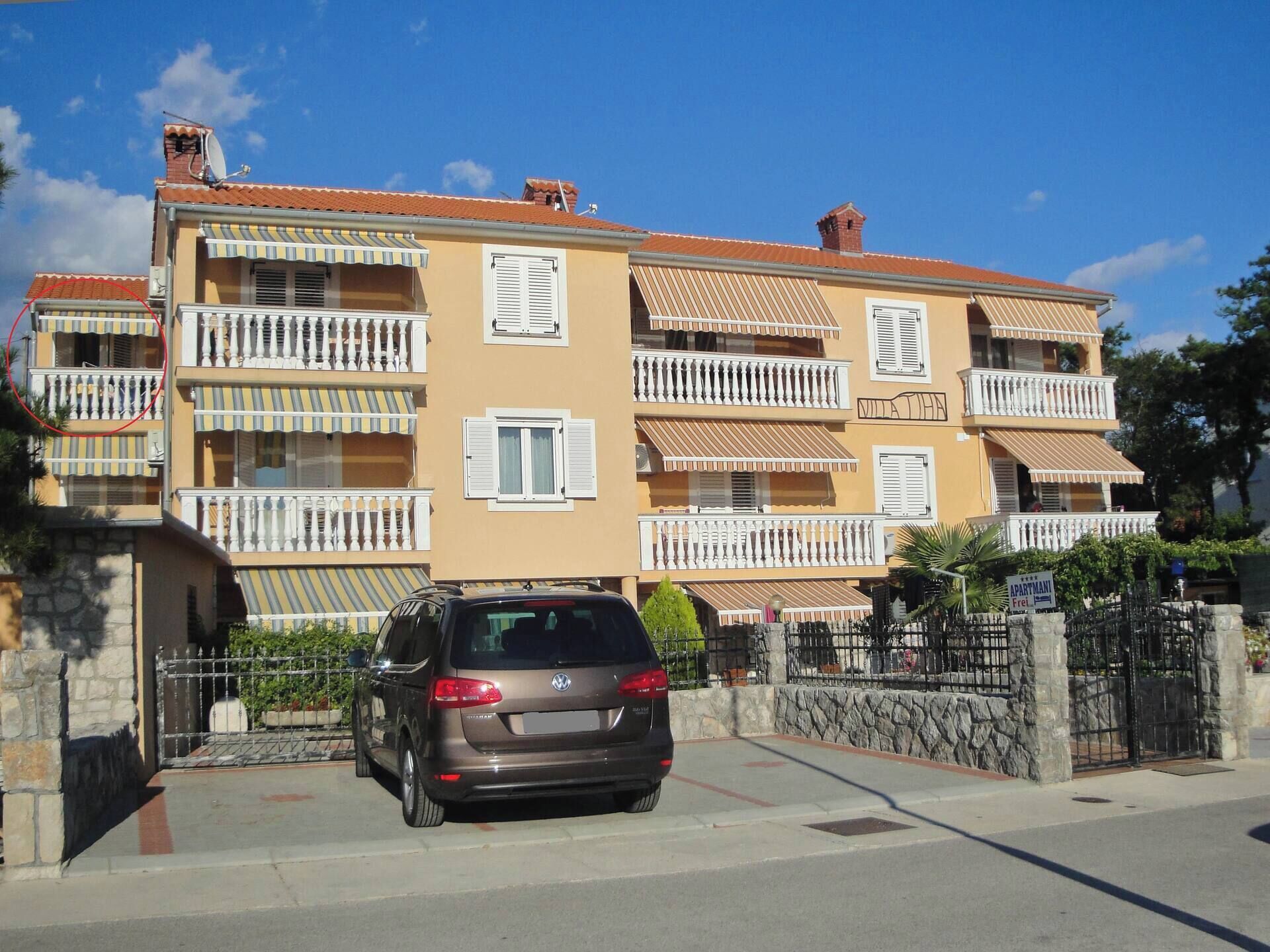 A multi-story residential building with balconies, a car parked in front, and a stone wall with a gate.
