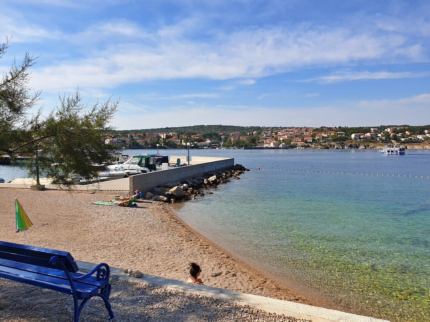 Una playa de guijarros con un banco, un pequeño refugio y barcos al fondo.