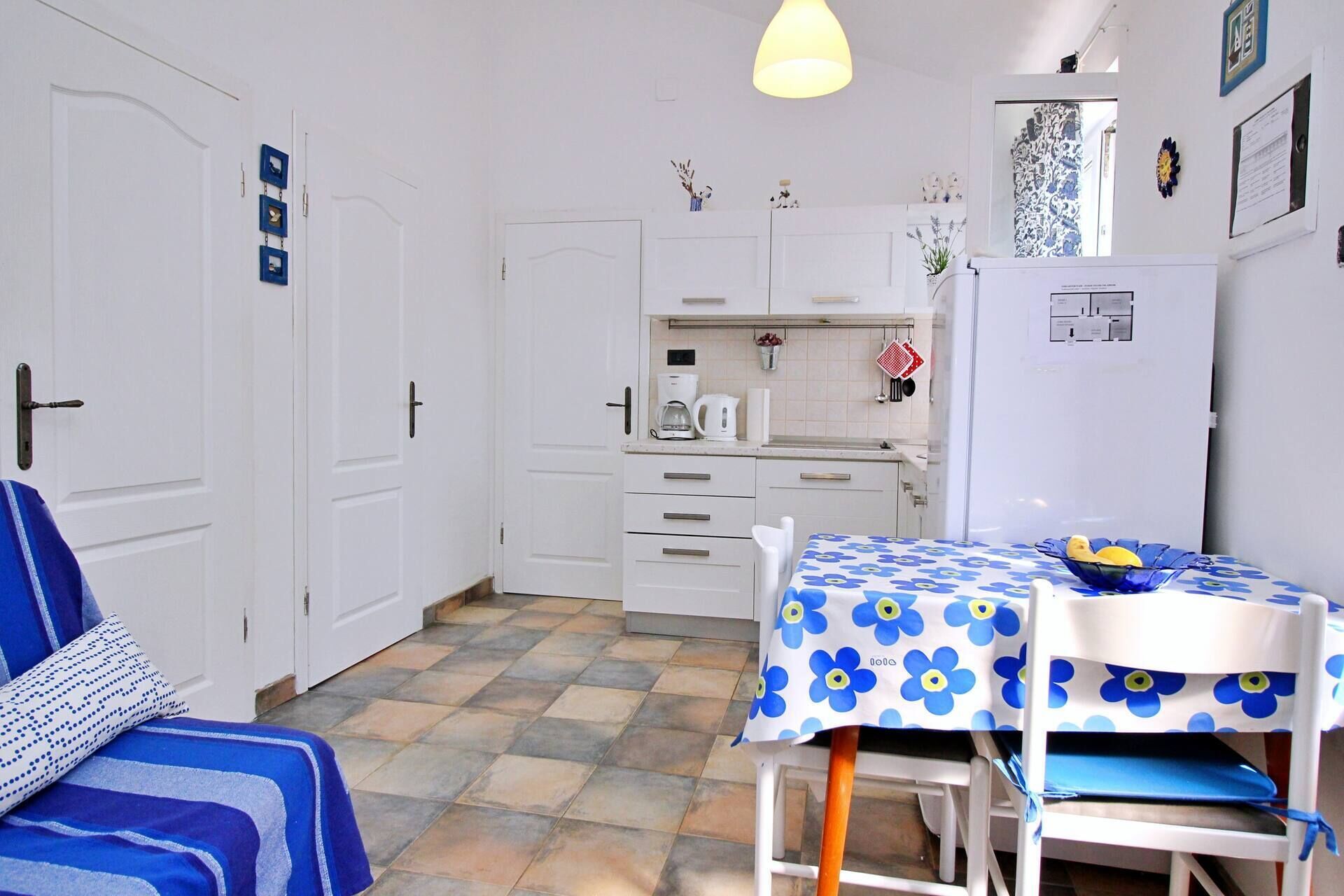 Een kleine keuken met witte kastjes, een koffiezetapparaat en een koelkast. Er staat een eettafel met een blauwe tafelkleed met bloemenprint en een schaal met fruit.