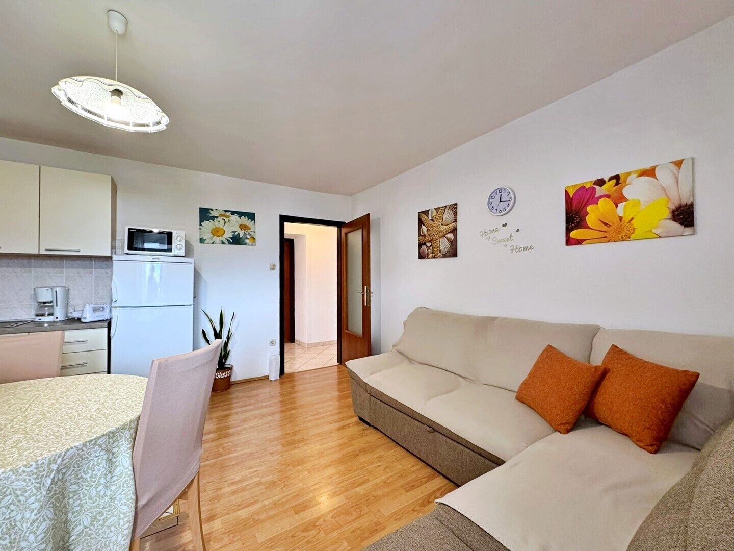 A modern apartment with a kitchenette, a dining area, a living room with a sofa and cushions, and a hallway leading to another room.