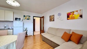 A modern apartment with a kitchenette, a dining area, a living room with a sofa and cushions, and a hallway leading to another room. - Apartment Car - 90 m from the sea, private terrace, parking, BBQ (Šilo)