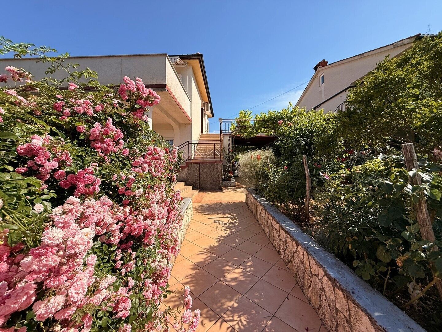 Un sendero que conduce a un edificio con un muro de piedra a un lado y un jardín con flores rosadas.