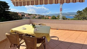 A terrace with a table and chairs, a striped awning, and a view of a coastal town and mountains. - Apartment Dragica - sea view, 350 m from sea, balcony, free parking, BBQ (Šilo)