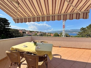 Una terraza con mesa y sillas, un toldo a rayas y vistas a un pueblo costero y montañas.