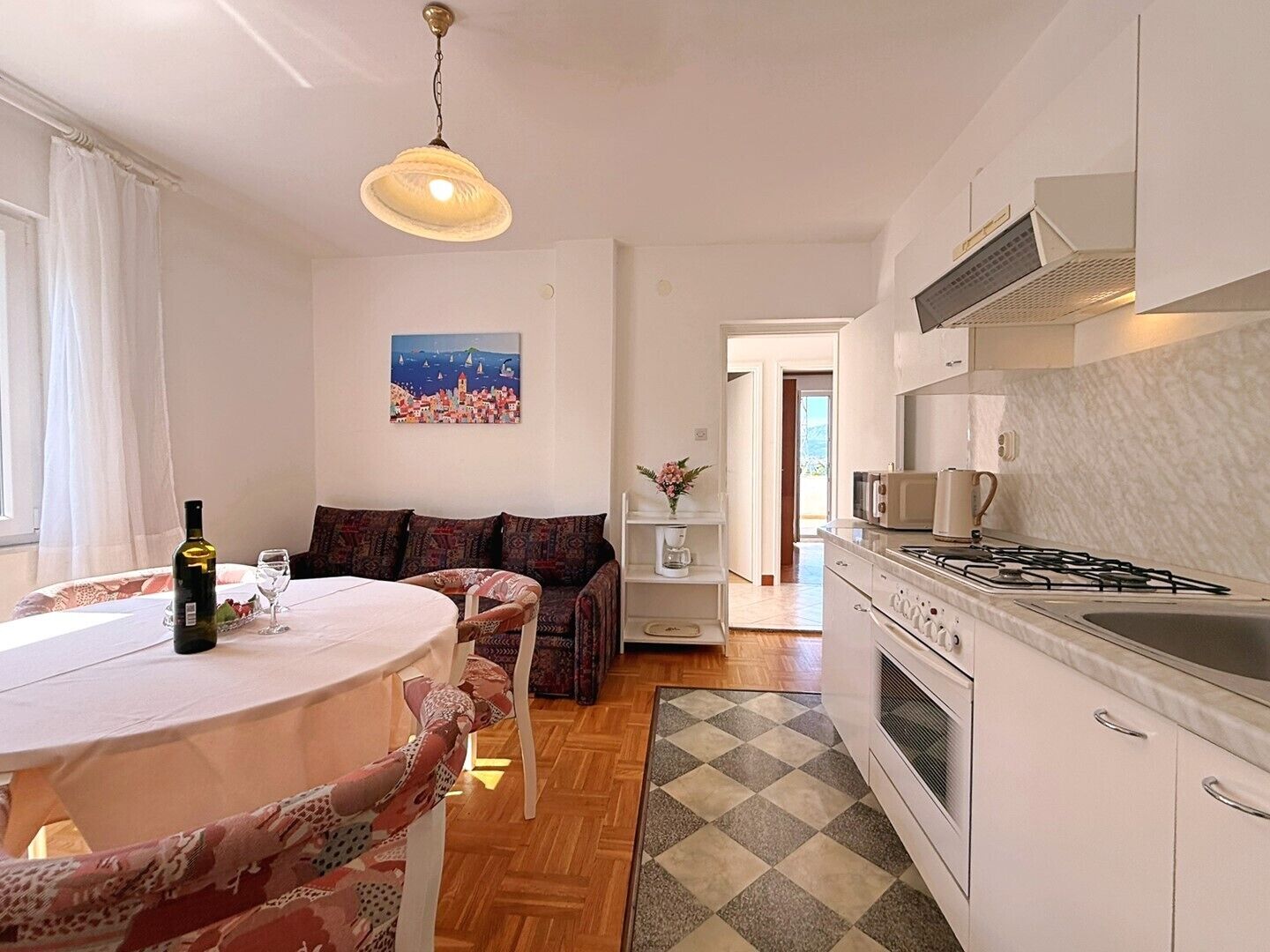 A modern kitchen with a gas stove, microwave, and white cabinets. There is a dining area with a table and chairs, and a sofa with patterned cushions. A painting hangs on the wall, and a window provides natural light.