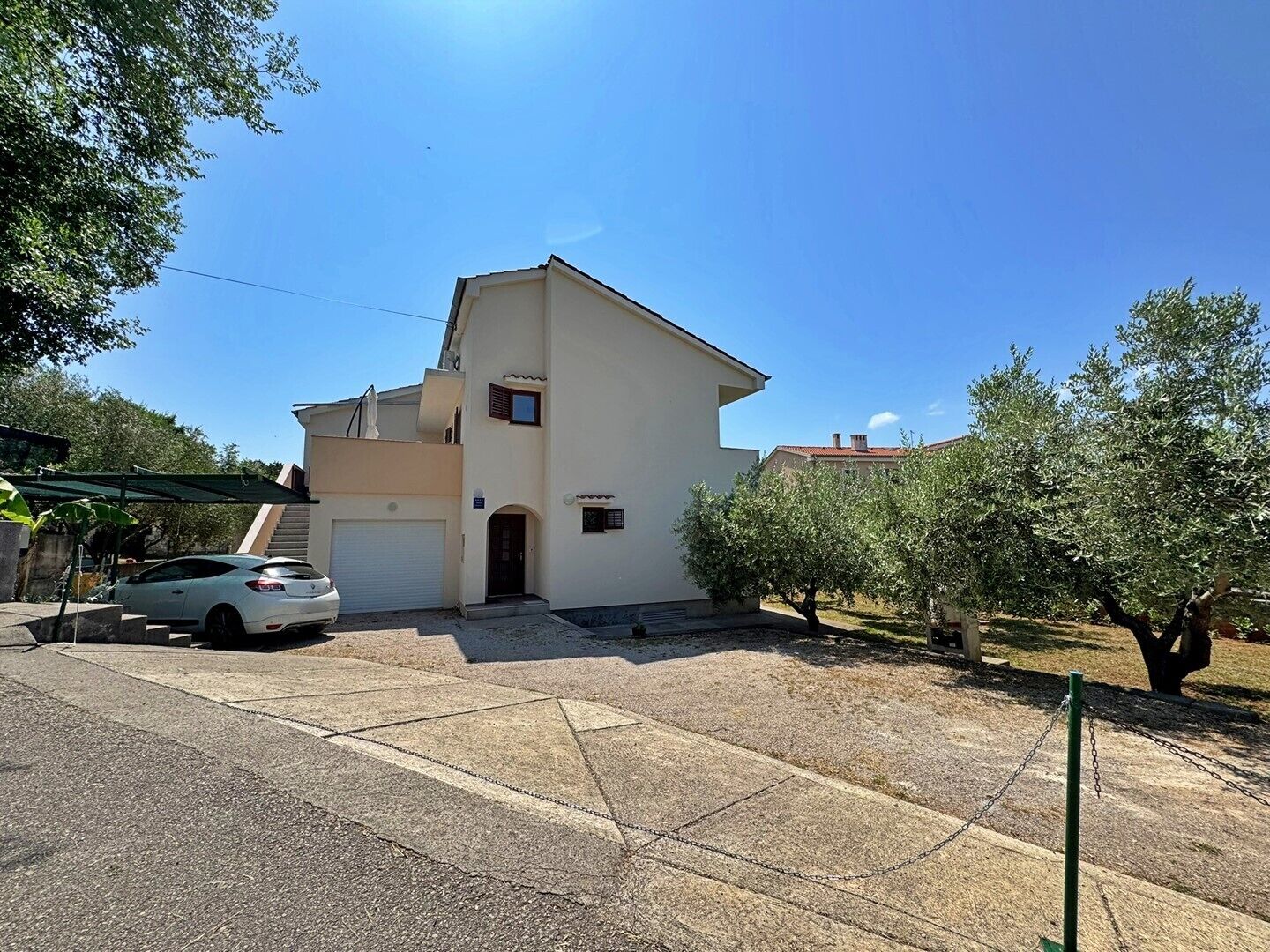 A two-story house with a white exterior, a garage, and a small garden with olive trees.